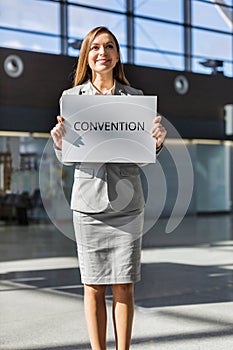 Portrait of young attractive businesswoman standing while holding white board with CONVENTION signage in arrival area at airport