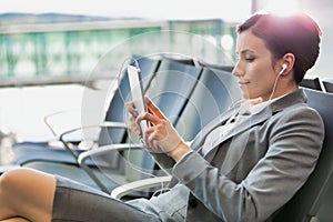Portrait of young attractive businesswoman using digital tablet with earphones on while waiting for boarding in airport with lens