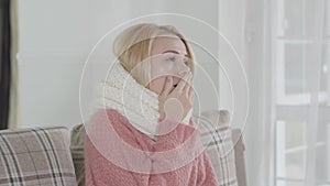 Portrait of young attractive Caucasian woman in white scarf shivering. Blond girl sitting at home on sofa and sneezing