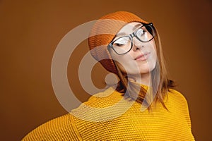 Portrait of a young attractive Caucasian woman in a yellow sweater and orange hat glasses on a brown background. Winter