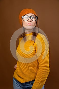 Portrait of a young attractive Caucasian woman in a yellow sweater and orange hat glasses on a brown background. Winter