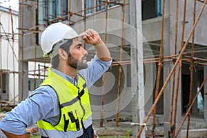 Portrait of young attractive construction man smiling bearded in vest with white helmet standing on construction site. Home