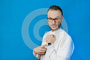 Portrait of young attractive european businessman in white shirt, glasses and beard pointing with hand at his smartwatch on blue