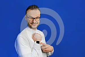 Portrait of young attractive european businessman in white shirt, glasses and beard pointing with hand at his smartwatch on blue