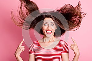 Portrait of young attractive excited crazy smiling girl point finger at flying hairstyle isolated on pink color