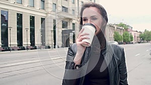 Portrait of a young attractive girl in a black leather jacket. standing in the cold wind and drinking coffee from a paper Cup. cit