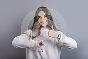 Portrait of a young attractive girl with dark long hair shows thumb up or down, good or bad on a gray studio background. Place for