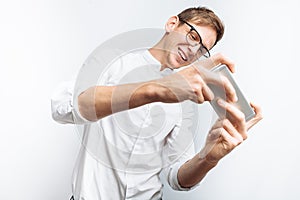 Portrait of young attractive guy in glasses playing games on phone, in white shirt isolated on white background, for advertising,