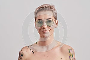 Portrait of a young attractive half naked tattooed woman wearing sunglasses and looking at camera isolated over light