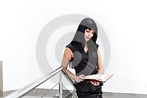 Portrait of Young Attractive happy brunette woman dressed in black business suit working with a notebook, standing in a office,