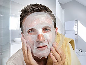 Portrait of young attractive and happy gay man in bathroom using beauty paper facial mask touching his face looking to the mirror