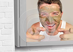 Portrait of young attractive and happy man laughing at himself in the mirror with green cream on his face applying facial mask an