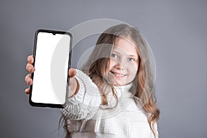 Portrait of a young attractive little girl with blond long flowing hair in a white sweater hold shows a mobile phone with a white