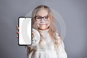 Portrait of a young attractive little girl with blond long flowing hair in a white sweater hold shows a mobile phone with a white