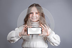 Portrait of a young attractive little girl with blond long flowing hair in a white sweater hold shows a mobile phone with a white