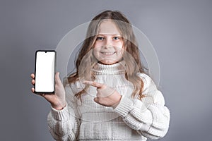 Portrait of a young attractive little girl with blond long flowing hair in a white sweater hold shows a mobile phone with a white
