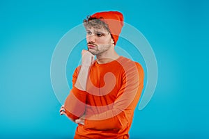 Portrait of young attractive man in orange hat and t-shirt stands in thinkful position on blue background