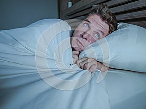 Portrait of young attractive scared man in fear and panic suffering horror nightmare covering face with blanket sleepless at night