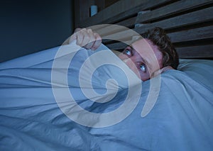 Portrait of young attractive scared man in fear and panic suffering horror nightmare covering face with blanket sleepless at night