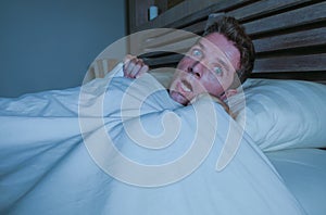 Portrait of young attractive scared man in fear and panic suffering horror nightmare waking up suddenly at night lying on bed in d