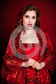 Portrait of a young, attractive vampire woman in a red rococo dress posing  against a dark background with red backlights