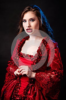 Portrait of a young, attractive vampire woman in a red rococo dress posing isolated against a dark background with blue backlights