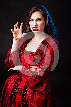 Portrait of a young, attractive vampire woman in a red rococo dress posing isolated against a dark background with blue backlights