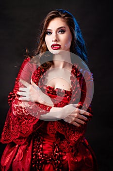 Portrait of a young, attractive vampire woman in a red rococo dress posing isolated against a dark background with blue backlights