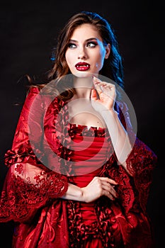 Portrait of a young, attractive vampire woman in a red rococo dress posing isolated against a dark background with blue backlights