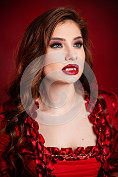 Portrait of a young, attractive vampire woman in a red rococo dress posing isolated against a dark background with red backlights