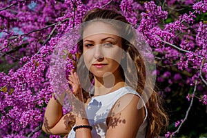 Portrait of young attractive woman with brown hair in white dress in blossom Judas tree. Beautiful girl poses gently in bloom tree