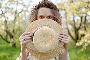 Portrait of a young attractive woman with curly hair hiding behind a wicker hat in a green garden. Spring mood