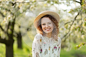 Portrait young attractive woman with curly hair in stylish wicker hat enjoys blooming green garden in spring day