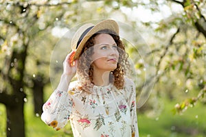 Portrait young attractive woman with curly hair in stylish wicker hat enjoys blooming green garden in spring day