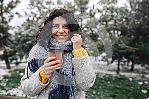 Portrait of a young attractive woman holding coffee at a snowy winter park. Female enjoys hot drink on a cold frosty winter day.