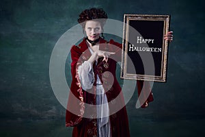 Portrait of young attractive woman in image of vampire holding picture frame  over dark green background. Horror