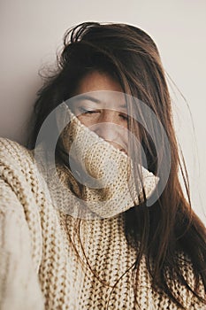 Portrait of young attractive woman with no makeup in knitted sweater smiling in soft light in room. Stylish hipster girl taking s