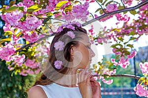 Portrait of young attractive woman with pink flowers in her hair is in white dress poses tender in blossom sakura tree in the park