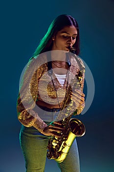 Portrait of young attractive woman, saxophonist performing jazz melodies in neon light against gradient background.