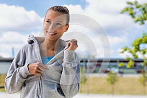 Portrait of young attractive woman smiling while standing and zipping her jacket in park