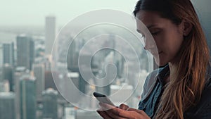 Portrait of young attractive woman standing near the window in skyscraper in Chicago, USA and using smartphone.