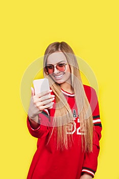 Portrait of a young attractive woman in sunglasses making selfie photo on smartphone isolated on a yellow background
