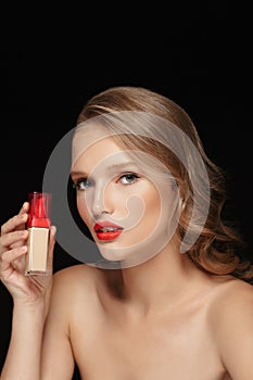 Portrait of young attractive woman with wavy hair and red lips holding concealer cream in hand while thoughtfully