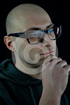 Portrait of a young bald man in black eyeglasses holding his chin with his hand while thinking looking away