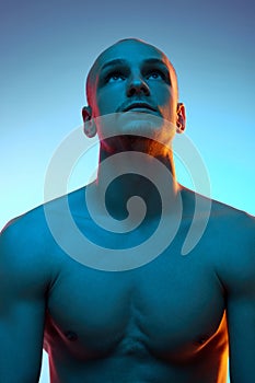 Portrait of young bald man in his 20s with muscular shirtless body sitting and looking upwards against blue studio