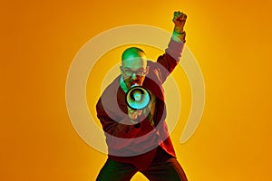 Portrait of young bald man with moustache in suit emotionally shouting in megaphone against orange studio background in