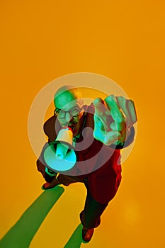 Portrait of young bald man with moustache in suit emotionally shouting in megaphone against orange studio background in