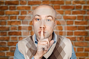 Portrait young bald man showing silence gesture with his finger in the mouth on brick wall background