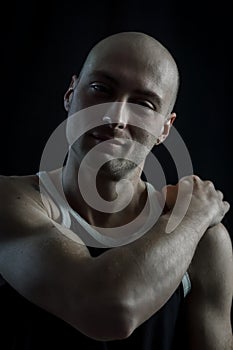 Portrait of a young bald man in an undershirt massaging his shoulder with his hand
