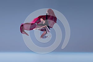 Portrait of young ballerina dancing with deep red fabric isolated over blue grey studio background. Graceful performance